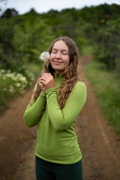 Joyful curly haired woman embracing a dandelion on a lush natural trail, expressing happiness and a peaceful connection with nature.