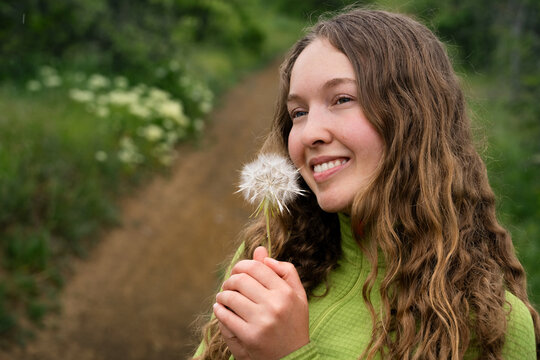 Natural close up portrait of a smiling curly haired woman with a dandelion in a lush spring landscape, expressing happiness and tranquility.