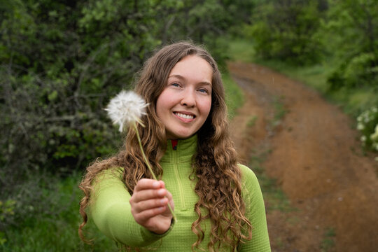 Happy woman with curly hair holding a dandelion flower toward the camera, surrounded by lush spring greenery.
