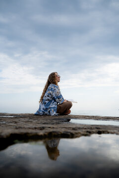 Relaxed young woman with curly hair reading on a coastal pier, surrounded by calm water, open cloudy sky and a beautiful reflection in the foreground. Casual lifestyle, technology and social detox.
