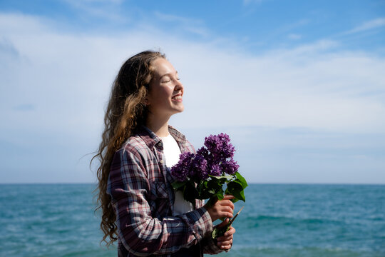 Joyful candid woman stands by the coast enjoying spring nature, blue sky, and fresh sea air with fresh lilac blossoms. Natural portrait embodies natural beauty, happiness, love, and enjoyment of life