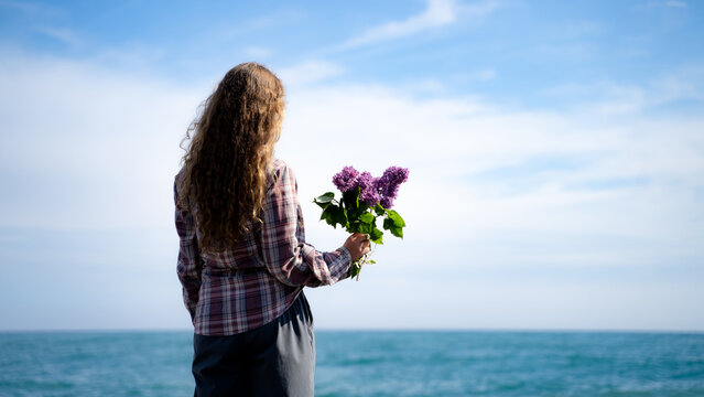 Back view of a long-haired, curly woman wearing a casual outfit, holding fresh lilac flowers and gazing towards the sea while standing on the beach. The coastal horizon on a sunny spring day.