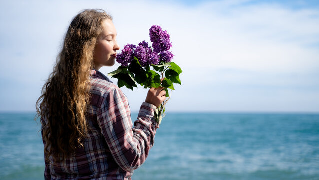 Beautiful woman enjoying the scent of fresh lilac blossoms while standing on a peaceful coastline, surrounded by blue sea and sky. A girl is enjoying the arrival of spring.