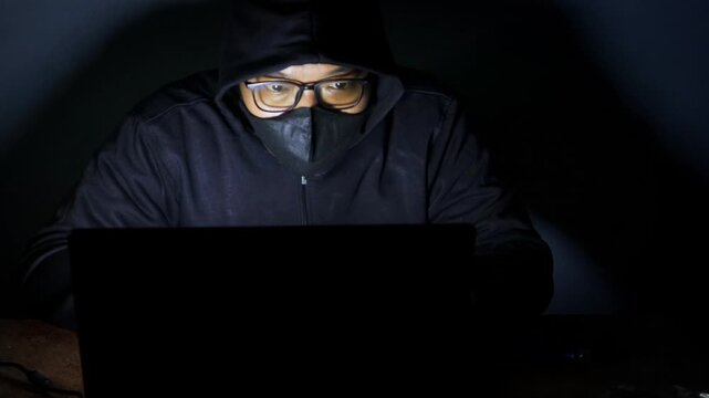 Male hacker wearing a hood, glasses, and face mask while working on a laptop in a dark room, accessing an online website or security system.