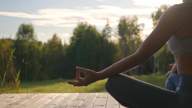 Close-up of Caucasian female arm and hand forming a meditation mudra with thumb and index finger touching while meditating outdoors overlooking a peaceful meadow and forest in soft morning light.