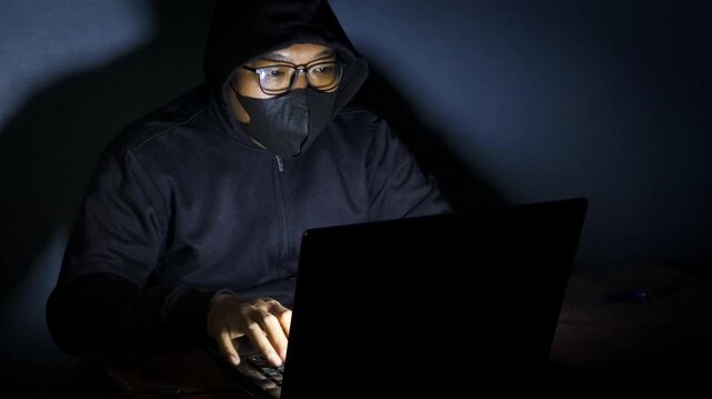 Male hacker wearing a hood, glasses, and face mask while working on a laptop in a dark room, accessing an online website or security system.
