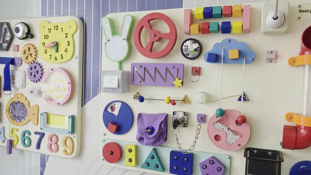 A static shot of two colorful educational sensory boards, or busy boards, mounted on a white and blue patterned wall inside a children's room or playroom.