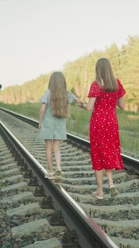Two young women hold hands as they stroll down parallel railroad tracks. One girl balances her foot on top of each metal tie while wearing striped shirt and denim overalls other wears red