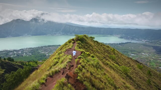 Man running along a grassy mountain ridge trail on Mount Batur, with Lake Batur and the surrounding volcanic caldera landscape visible under a cloudy sky in Bali, Indonesia. Aerial view drone flight