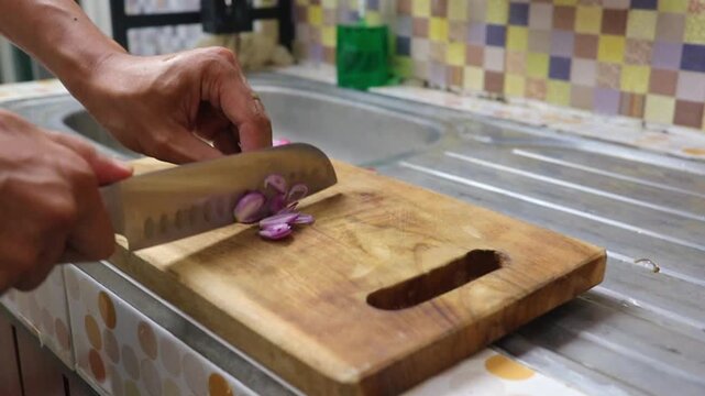 People hands slicing fresh shallots using a sharp knife on a wooden cutting board in a home kitchen