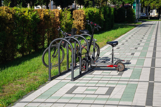 Electric scooter and bicycle parked at metal stand on tiled sidewalk of urban park zone promoting green city infrastructure