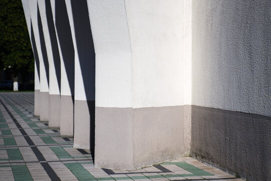 Modern building concrete facade with rhythmic pillars casting deep geometric shadows on tiled public sidewalk pavement with copy space