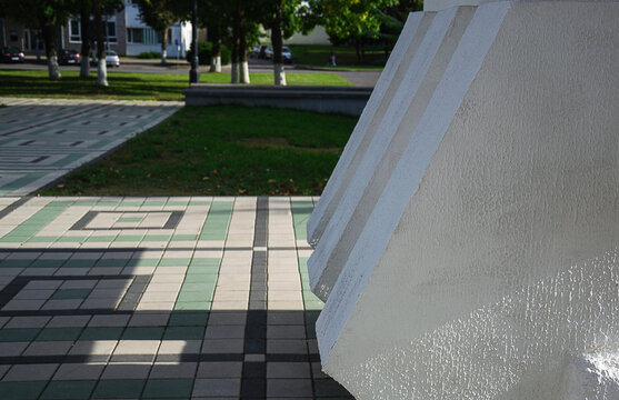 Massive white concrete structural element stands next to patterned green and grey tiled pavement of outdoor public plaza near city park zone with copy space