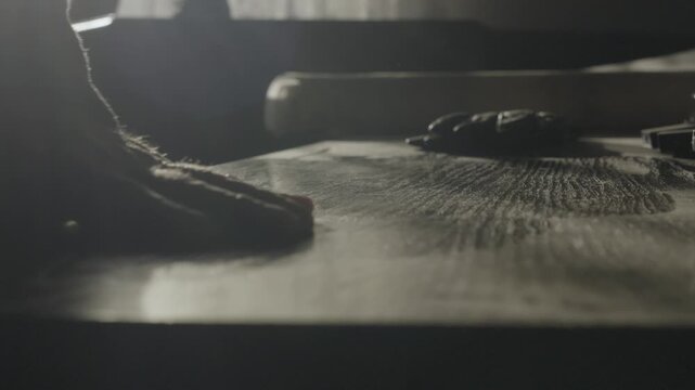 Close up of a hand slowly moving across a wooden tabletop, feeling its surface texture under dramatic window lighting, creating a tactile atmospheric concept.