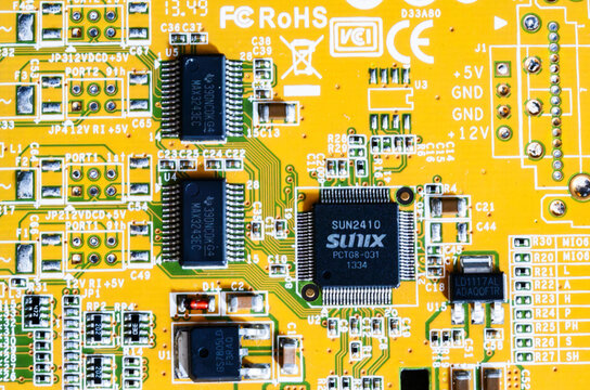 Detailed close-up of a yellow printed circuit board featuring branded microchips, capacitors, and compliance logos.