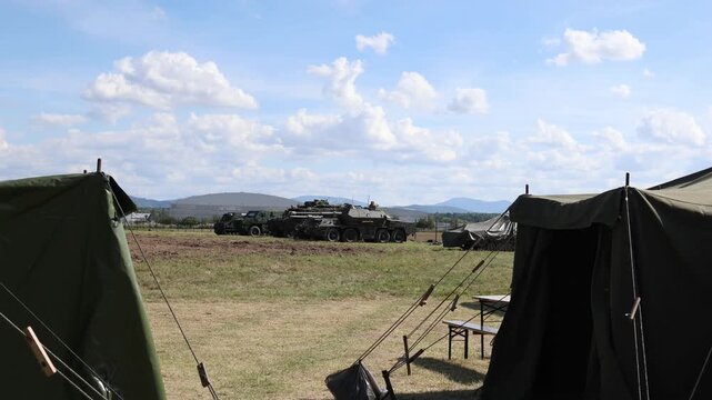 Military field camp with Pandur II 8x8 CZ armored personnel carrier and support vehicles parked between tents during an outdoor defense equipment display in daylight view