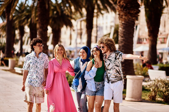 Happy Diverse Friends Walking Together Along Sunny Palm-Tree Lined Promenade On Summer Vacation
