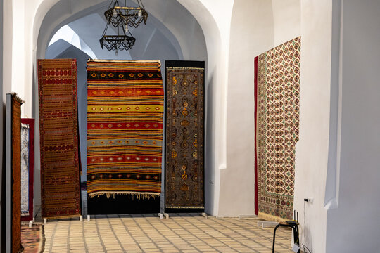 Bukhara, Uzbekistan. Colorful woven rugs and patterned textiles hang beneath white arched architecture, while geometric designs, hanging lanterns and chair define the interior display