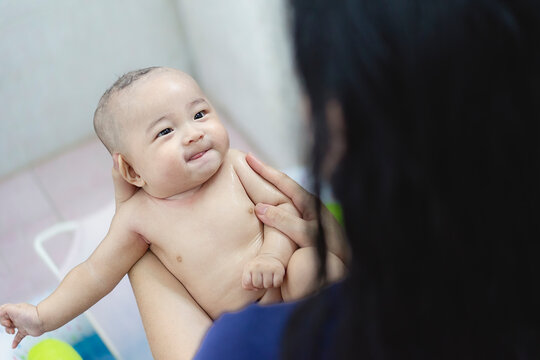 Baby bath time and infant hygiene concept, caring mother holding wet Asian baby smiling with tongue out in bathtub, representing maternal care, gentle parenting, and newborn wellness.