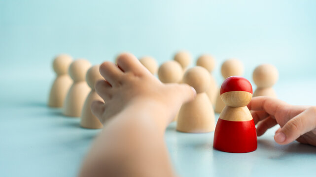 Child choosing leader and talent selection concept, kid hands reaching toward red wooden peg doll standing before crowd of natural figures on blue background, symbolizing decision, and role model.