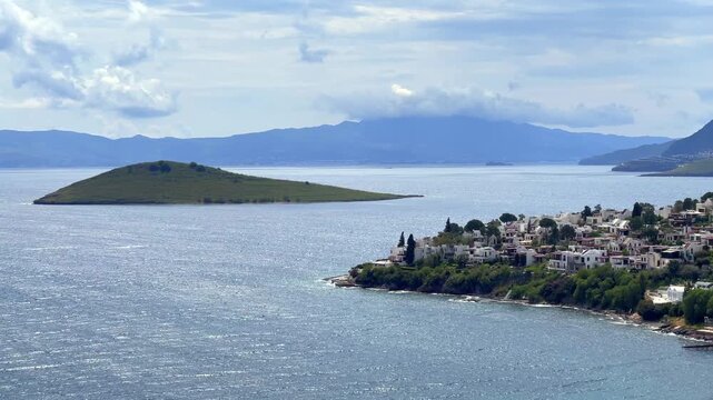 A elevated scenic view of Bitez in Bodrum, Turkey, showing white coastal houses, blue sea waters, an island, and mountain ridges under a cloudy sky.