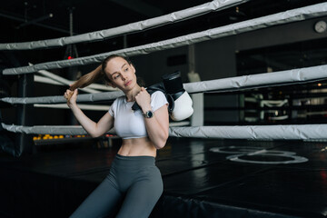 Obraz premium Female athlete holding boxing gloves and adjusting hair, taking break inside boxing ring after challenging workout, embodying dedication and strength in fitness journey.