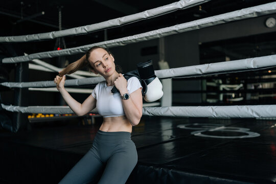 Female athlete holding boxing gloves and adjusting hair, taking break inside boxing ring after challenging workout, embodying dedication and strength in fitness journey.
