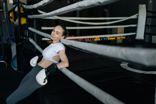 Woman athlete wearing boxing gloves leaning on ropes of boxing ring, taking break from training in gym environment. Concept of on fitness journey and strength, healthy sports life.