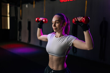 Obraz premium Focused woman with muscular body training with dumbbells in fitness club. Low key lighting with purple and red accents creates motivated atmosphere for strength training and bodybuilding.