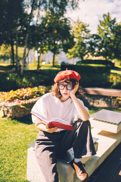 Young student in red beret looks bored reading a book in a park