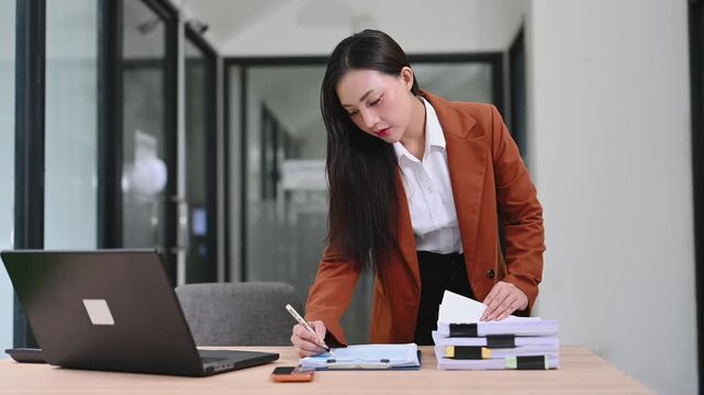 Smiling Asian businesswoman in a stylish blazer elegantly signs corporate documents at her modern office desk. A true portrait of professional success and focused administration.