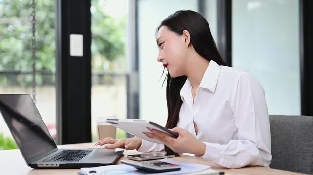 Smiling Asian businesswoman multitasking with a digital tablet and laptop at a modern office desk. A confident professional working online