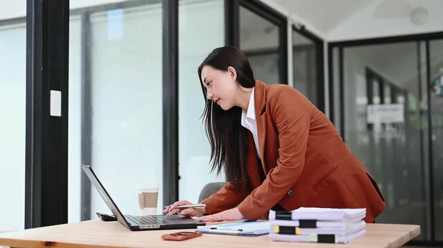 Smiling Asian businesswoman in a stylish blazer elegantly signs corporate documents at her modern office desk. A true portrait of professional success and focused administration.