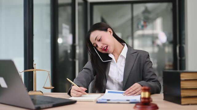 Asian female attorney multitasking on a call while reviewing contracts. A portrait of professional success at a desk with a gavel and scales of justice.