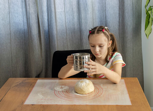 A little girl of european appearance sits at a wooden table and uses a metal sifter to dust flour over a ball of fresh dough on a silicone mat while baking at home. Home cooking concept.