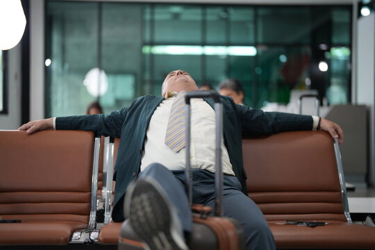 A senior businessman stretched across the seats sprawls across the bench during a long delay on the seating row of an airport gate, with the terminal quiet around him