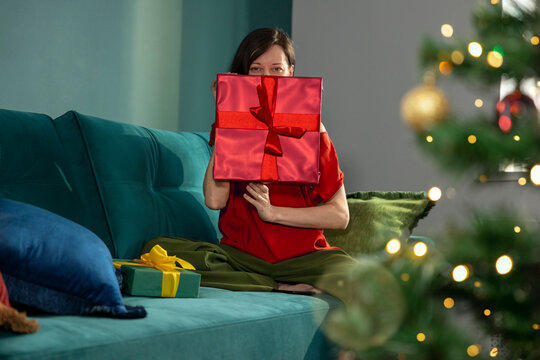 Woman hides her face behind a red Christmas gift on a sofa