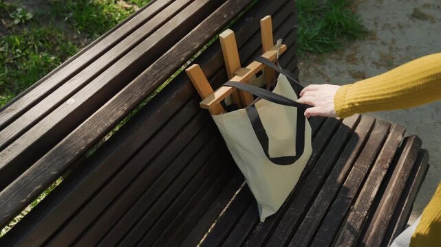 Person in yellow sweater places and organizes painting supplies into bag beside empty wooden bench outdoors. scene includes visible materials like brushes wood fabric bags grassy areas
