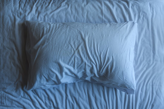 Single white pillow on wrinkled bed sheets under soft cool blue bedroom light