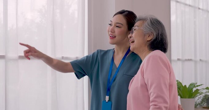 Female Asian caregiver walks beside senior Asian woman, keeps arm around back and points ahead during interaction in assisted living center, promoting companionship, confidence and wellbeing.
