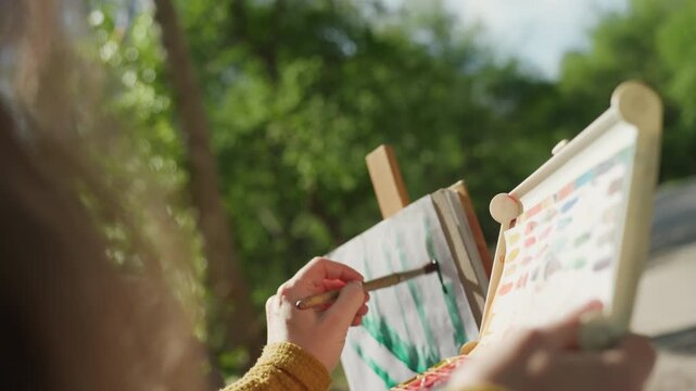 Close up view of hands holding paintbrushes selecting colors from portable color palette outdoors. artist applies green brushstrokes to canvas on easel in natural light