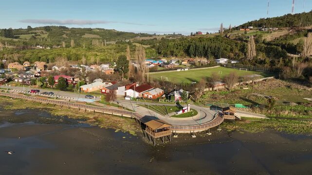A wooden coastal walkway curves along the tidal flats of Curaco de V&eacute;lez, a lookout deck resting at its end as houses, a soccer field, and forested hills frame this quiet Chilo&eacute; town.