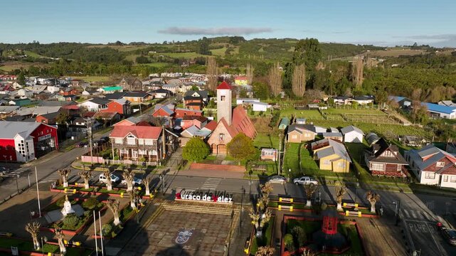 Warm evening light spreads across the town square of Curaco de V&eacute;lez, its stone church and colorful traditional houses framed by quiet streets and rolling farmland in Chilo&eacute;, Chile.