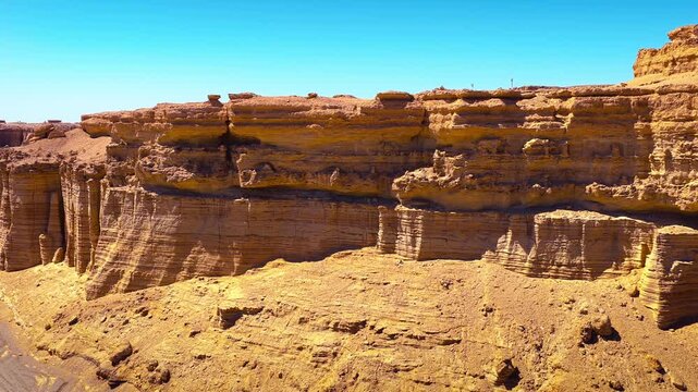 Layered yardang landform cliffs rise above a desert track in Dahaidao, Xinjiang, captured under a bright blue sky.