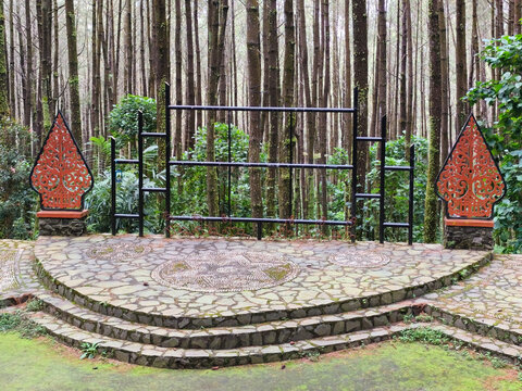 ​An open air performance stage decorated with Javanese gunungan ornaments set inside a dense pine forest, representing local culture, traditional arts, eco tourism, and natural event venue design.