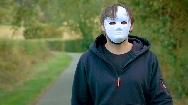 Close view of masked criminal approaching camera on lonely rural road. Anonymous murderer and killer wearing white mask and black hoodie. Maniac, rapist, violent crime, assault, victim and dangerous