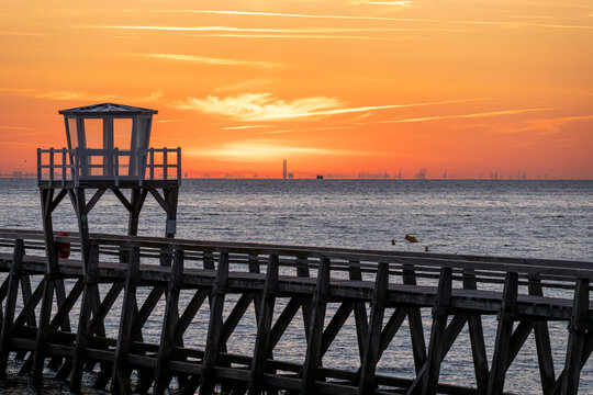 Luc-Sur-Mer, France - 08 08 2026:  View of a wooden pier in front of the sea, a colorful sunrise and port of Le Havre far away