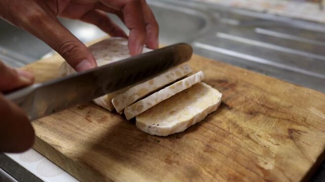 People hands slicing raw tempeh using a sharp knife on a wooden cutting board in a home kitchen.