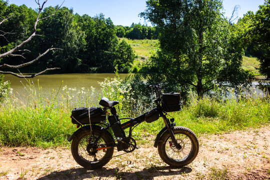 Modern foldable e-bike with fat tires parked on a dirt trail near a calm summer lake, perfect for eco-friendly adventure and weekend outdoor biking: Russia - July 17, 2026