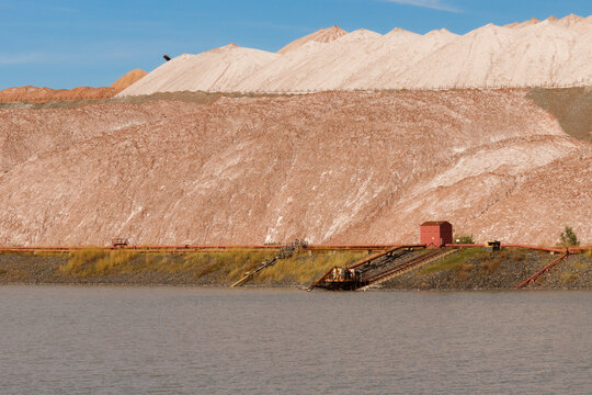 Multi layered brown and white salt slag heaps of a potash mining quarry rising behind a small technical water reservoir pond under a blue sky sunny day horizontal view